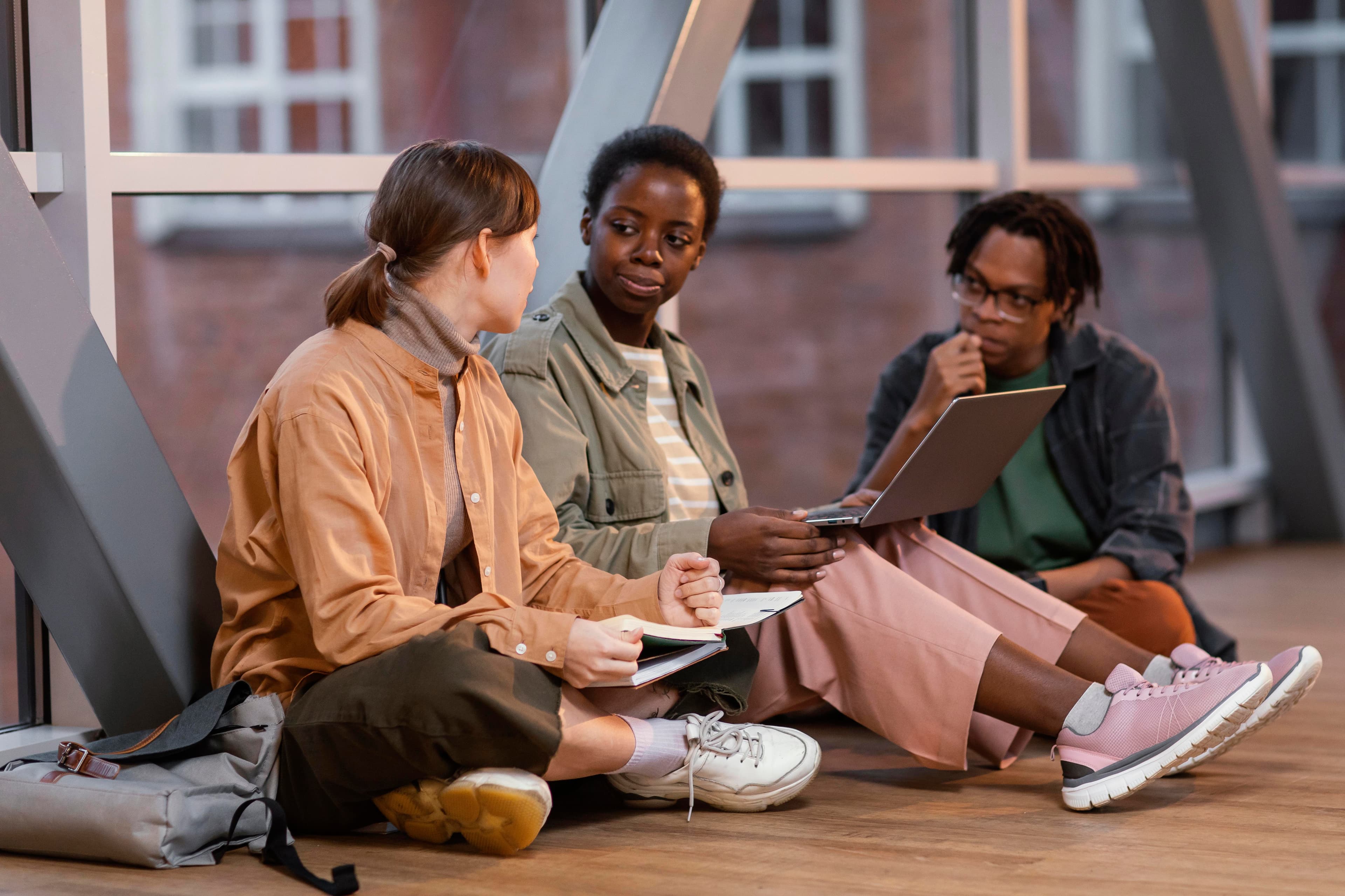 Three learners sitting together on a floor, collaborating on a project with a laptop and notebook.